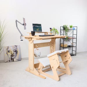 May include: A light-coloured wooden standing desk with a matching kneeling chair. The desk has a laptop, keyboard, and monitor arm. A black mug, headphones, and plants are also on the desk. A black metal shelf and artwork are in the background.