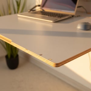 May include: A white desk surface with a visible layered plywood edge, featuring two small circular metal insets near the corner. The desk is positioned with a laptop and a computer mouse resting on its flat, smooth top surface.