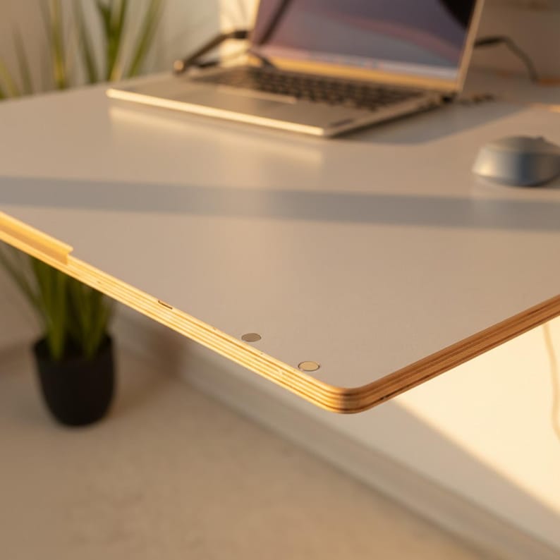May include: A white desk surface with a visible layered plywood edge, featuring two small circular metal insets near the corner. The desk is positioned with a laptop and a computer mouse resting on its flat, smooth top surface.
