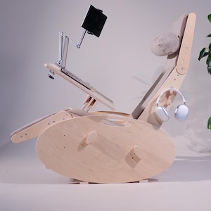 K&ouml;nnte beinhalten: Ein hellfarbiger Schaukelstuhl aus Holz mit eingebautem Laptop-St&auml;nder und Tablethalter. Der Stuhl hat eine Kopfst&uuml;tze und Armlehnen mit einem hellfarbigen Stoff. Ein wei&szlig;er Kopfh&ouml;rer h&auml;ngt an der Seite.