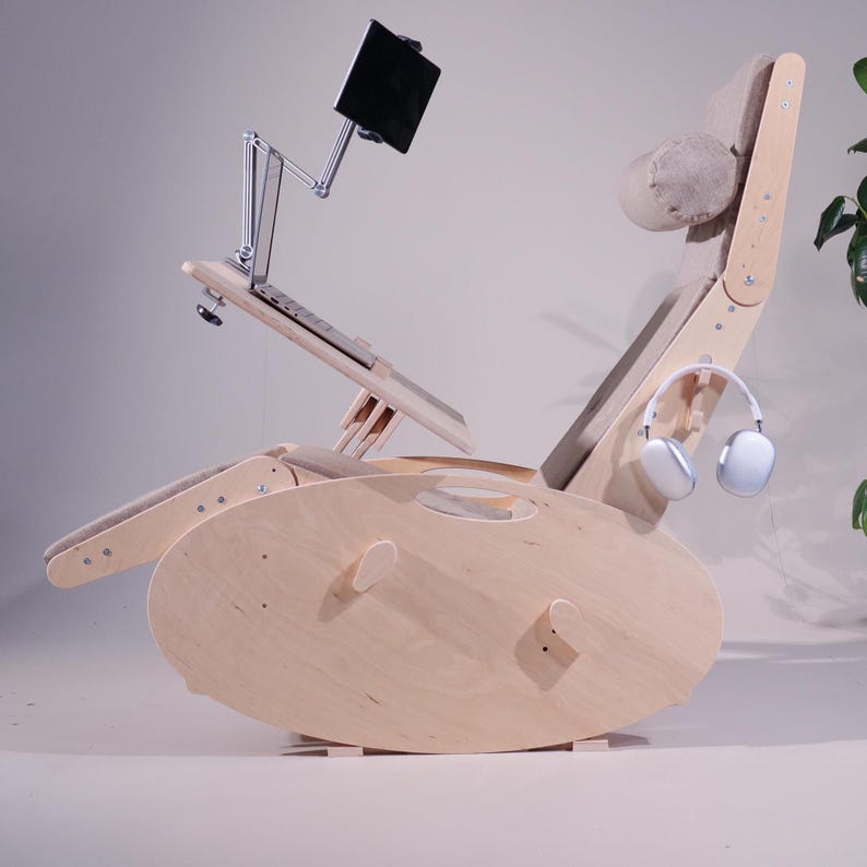K&ouml;nnte beinhalten: Ein hellfarbiger Schaukelstuhl aus Holz mit eingebautem Laptop-St&auml;nder und Tablethalter. Der Stuhl hat eine Kopfst&uuml;tze und Armlehnen mit einem hellfarbigen Stoff. Ein wei&szlig;er Kopfh&ouml;rer h&auml;ngt an der Seite.