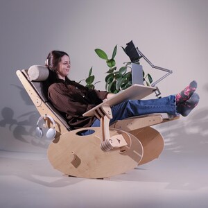 May include: A wooden rocking chair with a built-in desk and headrest. The chair has a natural wood finish and a gray fabric seat. A laptop and tablet are on the desk. Headphones hang on the side. A plant is in the background.