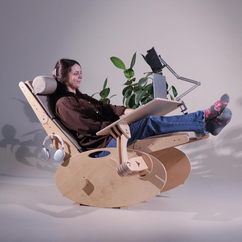 May include: A wooden rocking chair with a built-in desk and headrest. The chair has a natural wood finish and a gray fabric seat. A laptop and tablet are on the desk. Headphones hang on the side. A plant is in the background.