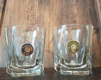 May include: Two clear glass tumblers with a gold-colored bullet casing embedded in the bottom of each glass. The bullet casings have the text "Remington 12 GA" and "Winchester 12 GA" on them.
