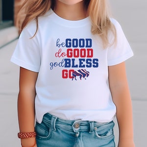 May include: A white t-shirt with a red, white, and blue graphic that says "be good do good god bless go". The graphic also includes a silhouette of a buffalo. The shirt is being worn by a young person wearing a pink hat and sunglasses.