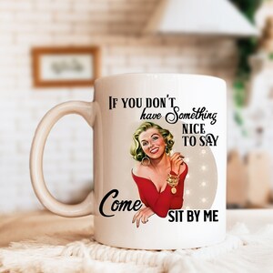 May include: White ceramic mug with a red and gold design. The mug features a vintage-style illustration of a woman with blonde hair and a red dress. The text on the mug reads "If you don't have something nice to say, come sit by me."