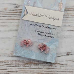 May include: A pair of rose gold glitter bow earrings on a blue and white card with the text "Hastrich Designs" and the website address "www.HastrichDesigns.com" and "www.etsy.com/shop/HastrichDesigns".