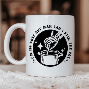 May include: White ceramic mug with a black circle design featuring a skeleton hand stirring a pot with the text "I'm no chef but man can I stir the pot."