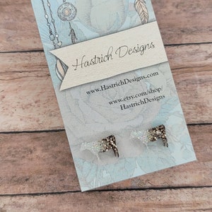 May include: A pair of cow-shaped earrings with glitter on a blue background. The earrings are on a card with the text "Hastrich Designs" and website information.