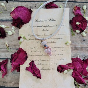 May include: A white leather cord necklace with a pink and red crystal pendant. The pendant is surrounded by dried rose petals and white flowers. The text on the card reads "Healing and Wellness" and describes the benefits of the crystal.