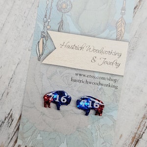 May include: A pair of red, white, and blue glitter buffalo earrings with the number "716" on each. The earrings are on a white card with the text "Hastrich Woodworking & Jewelry" and "www.etsy.com/shop/hastrichwoodworking".
