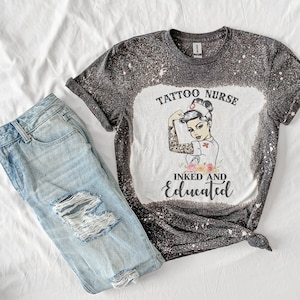 May include: A black and white graphic tee shirt with a distressed look. The shirt features a cartoon image of a female nurse with a tattoo on her arm. The text on the shirt reads "Tattoo Nurse Inked and Educated".