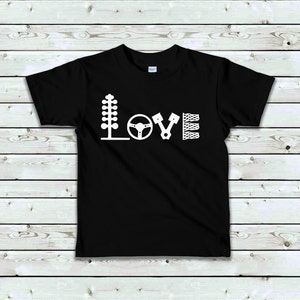 May include: Black t-shirt with a white graphic of the word "LOVE" spelled out using racing-themed icons. The icons include a Christmas tree, a steering wheel, a piston, and a chequered flag.