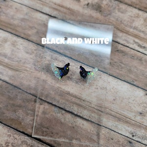May include: A pair of black and white glitter chicken earrings. The earrings are on a clear display card with the text "BLACK AND WHITE" printed on it.