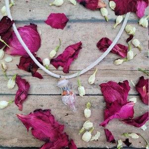 May include: A white cord necklace with a small glass bottle pendant filled with dried flowers and crystals. The necklace is lying on a wooden surface with dried rose petals and white flowers scattered around it.