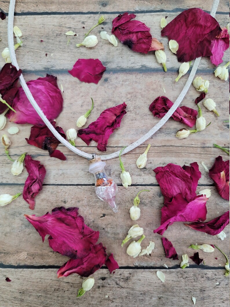 May include: A white cord necklace with a small glass bottle pendant filled with dried flowers and crystals. The necklace is lying on a wooden surface with dried rose petals and white flowers scattered around it.