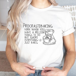 May include: White t-shirt with a black and white illustration of a stand mixer and the text "PROCRASTIBAKING VERB: WHEN YOU HAVE A MILLION THINGS TO DO BUT IGNORE EVERYTHING -&- JUST BAKE."