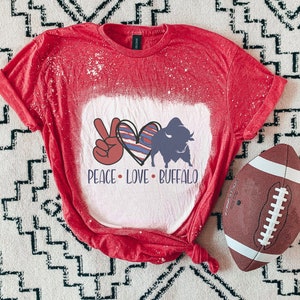 Könnte beinhalten: Rotes T-Shirt mit einem weiß gebleichten Design, das ein Peace-Zeichen, ein Herz mit rot-weiß-blauen Streifen und eine Büffel-Silhouette zeigt. Der Text "Peace Love Buffalo" ist unter dem Design gedruckt.
