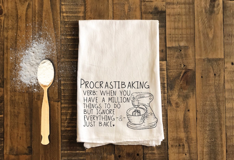 May include: White kitchen towel with black text that reads "PROCRASTIBAKING VERB: WHEN YOU HAVE A MILLION THINGS TO DO BUT IGNORE EVERYTHING-&- JUST BAKE." A black and white illustration of a stand mixer is to the right of the text.