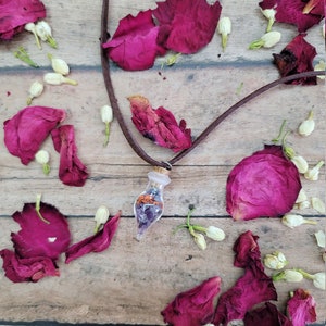 K&ouml;nnte beinhalten: Eine braune Lederkordelkette mit einem Glaskolben-Anh&auml;nger, gef&uuml;llt mit getrockneten Blumen und Kristallen. Die Kette ist umgeben von rosa Rosenbl&auml;ttern und wei&szlig;en Bl&uuml;tenknospen auf einer Holzoberfl&auml;che.