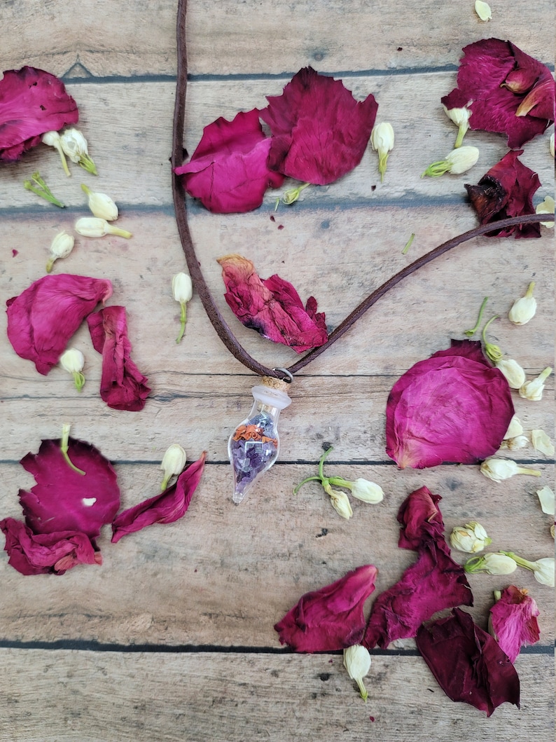 K&ouml;nnte beinhalten: Eine braune Lederkordelkette mit einem Glaskolben-Anh&auml;nger, gef&uuml;llt mit getrockneten Blumen und Kristallen. Die Kette ist umgeben von rosa Rosenbl&auml;ttern und wei&szlig;en Bl&uuml;tenknospen auf einer Holzoberfl&auml;che.
