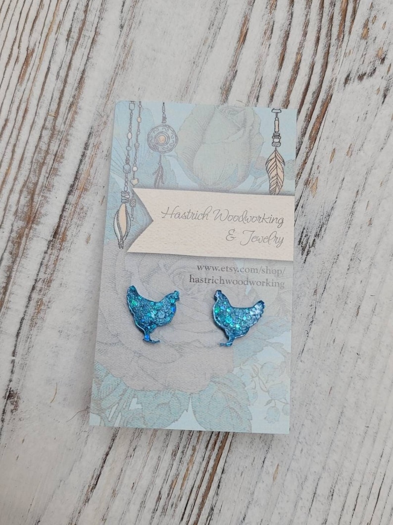 May include: A pair of blue glitter chicken earrings. The earrings are on a white card with the text "Hastrich Woodworking & Jewelry" and "www.etsy.com/shop/hastrichwoodworking".