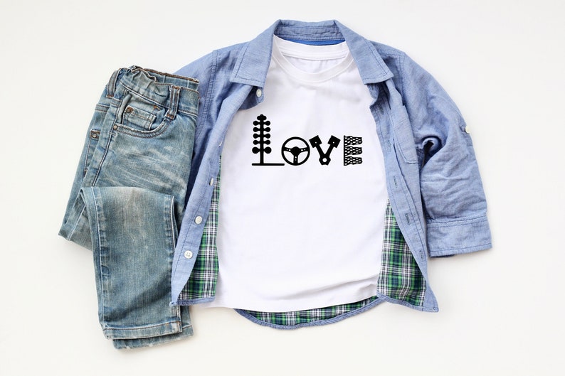 May include: White t-shirt featuring a black graphic of the word "LOVE" spelled out using racing-themed icons. The shirt is laid out on a white surface with a blue plaid shirt and blue jeans.
