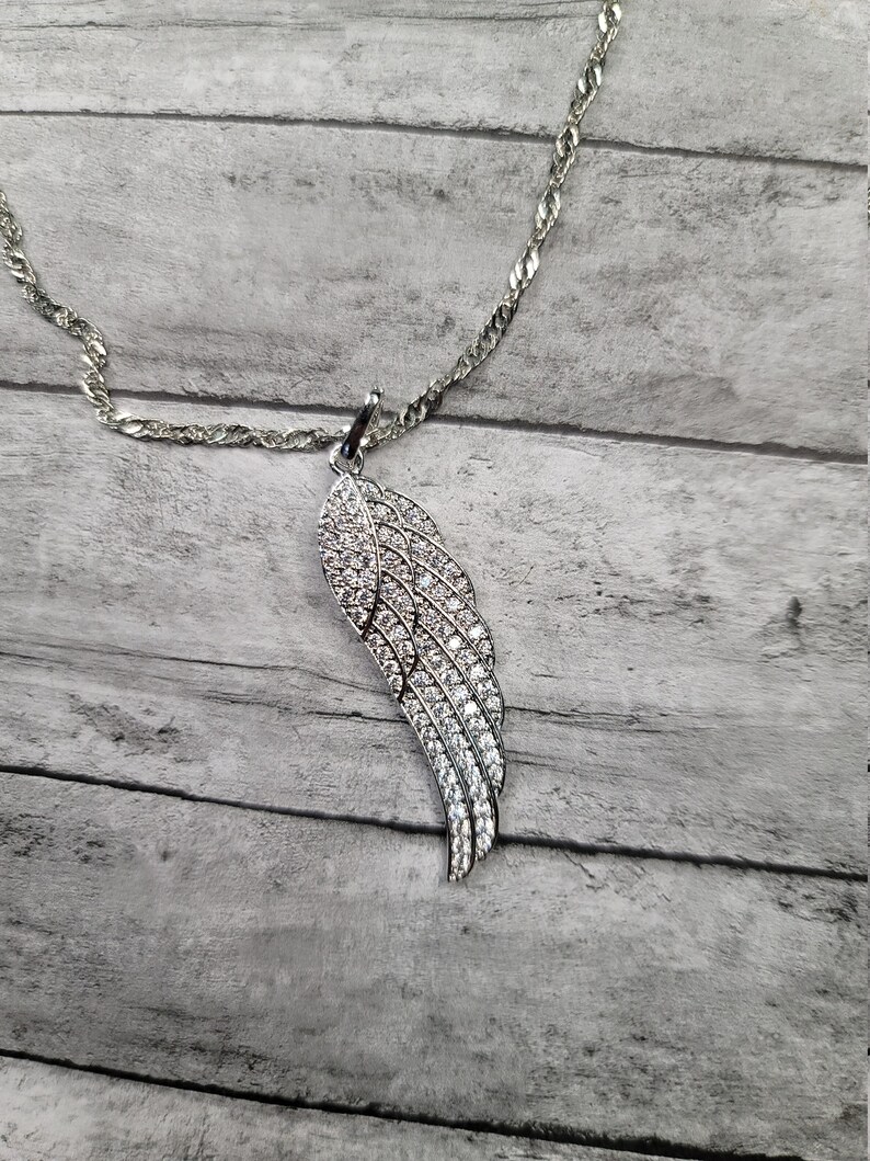 Pode incluir: Um colar de corrente de prata com um pingente de asa de prata incrustado com pedras de zirc&ocirc;nia c&uacute;bica transparentes.