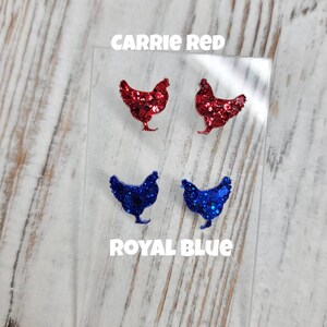 May include: Two pairs of glitter chicken earrings. One pair is red and the other is blue. The earrings are displayed on a clear plastic backing with the words "Carrie Red" and "Royal Blue" printed on it.