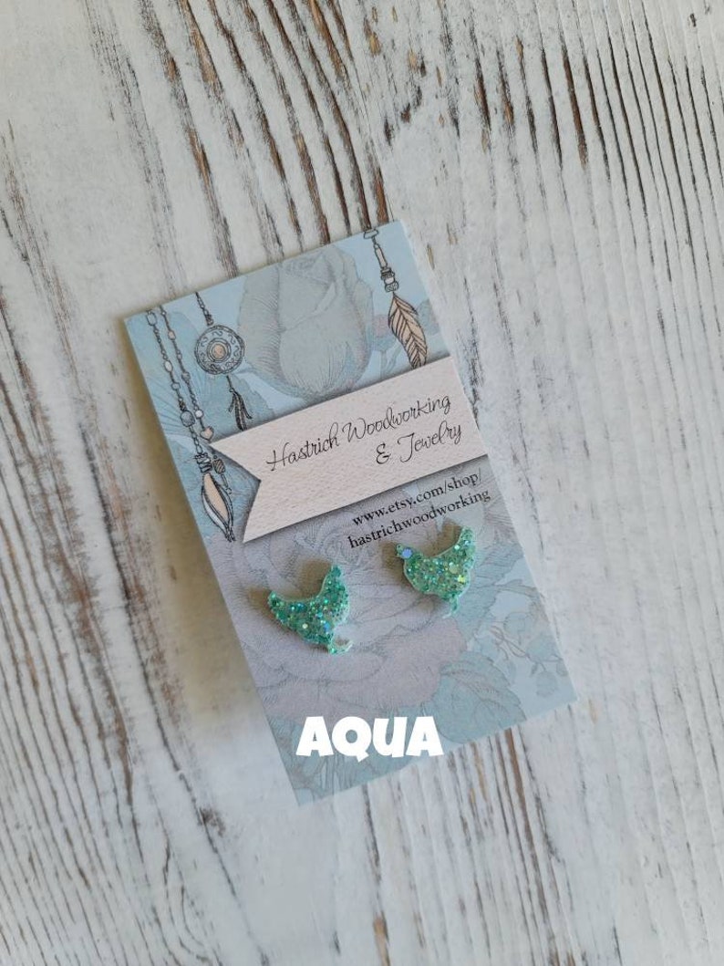 May include: A pair of aqua blue glitter bird earrings on a card with the text "Hastrich Woodworking & Jewelry" and "www.etsy.com/shop/hastrichwoodworking" and the word "AQUA" in large letters.