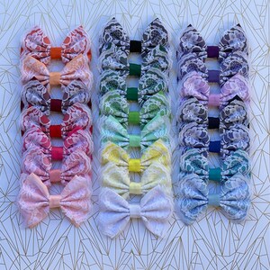 May include: A set of 10 handmade hair bows with a rainbow of colors. Each bow is made with lace fabric and has a different color. The bows are arranged in a row, with the colors transitioning from red to purple.