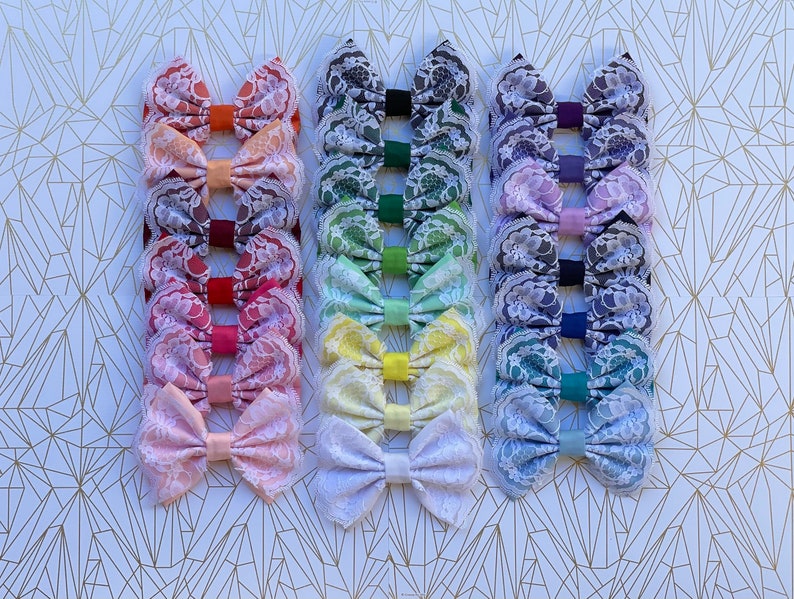 May include: A set of 10 handmade hair bows with a rainbow of colors. Each bow is made with lace fabric and has a different color. The bows are arranged in a row, with the colors transitioning from red to purple.