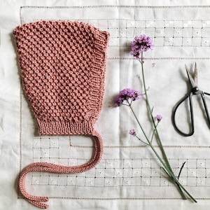 Puede incluir: Un gorro tejido en color rosa empolvado con un patrón de punto de gofre texturizado y lazos largos para la barbilla. Junto al gorro hay un tallo verde delgado con pequeños racimos de flores moradas y unas tijeras de manualidades negras.