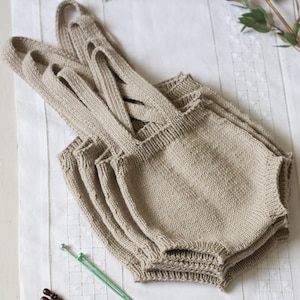 以下が含まれることがあります： 白い布の上に、ベージュのニット素材でできたベビーオーバーオールが3組重ねて置かれています。緑と茶色の編み針も一緒に置かれています。