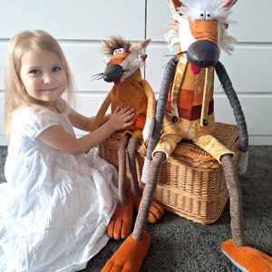 May include: Two whimsical fox dolls with long legs and orange feet sit on a wicker basket. The dolls have brown and orange fur, black noses, and long, thin whiskers. One doll wears a brown dress, and the other wears a patchwork outfit. A young girl in a white dress is in the image.