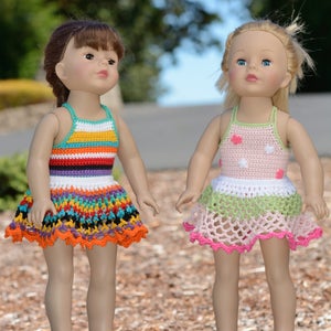 May include: Two dolls wearing crocheted dresses. The doll on the left is wearing a multi-colored striped dress with a ruffled hem. The doll on the right is wearing a pink and green dress with white polka dots and a ruffled hem.
