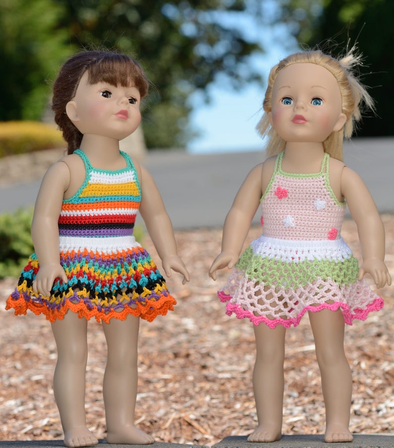 May include: Two dolls wearing crocheted dresses. The doll on the left is wearing a multi-colored striped dress with a ruffled hem. The doll on the right is wearing a pink and green dress with white polka dots and a ruffled hem.