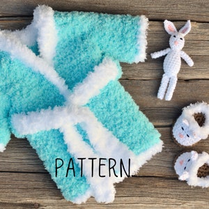 May include: A turquoise blue crocheted baby robe with a white fur trim and a white crocheted bunny toy. The robe has a tie belt and the word "PATTERN" is written on the front.
