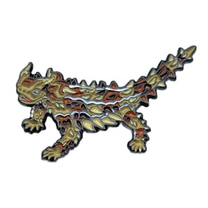 Pode incluir: Um broche de esmalte marrom e bege de um lagarto cornudo com uma faixa branca nas costas. O lagarto está em uma pose de corrida com a cauda levantada.
