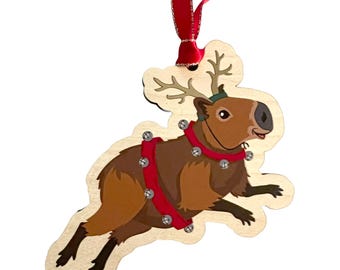 May include: Wooden Christmas ornament featuring a capybara wearing reindeer antlers and a red harness with bells. The ornament has a red ribbon for hanging. The capybara is brown with black feet and a green hat.