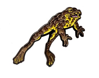 May include: An enamel pin featuring a frog in brown and yellow hues. The frog is shown with its legs outstretched. The pin has a shiny finish and is set against a white backdrop. The frog's mouth is open, revealing a pink interior.