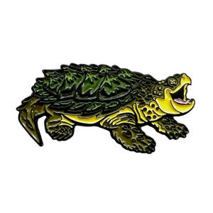 Pode incluir: Pin de esmalte detalhado retratando uma tartaruga mordedora. O pin apresenta uma carapaça texturizada verde escuro, uma cabeça amarela com a boca aberta e pernas marrons escuras. O design é delineado em preto.