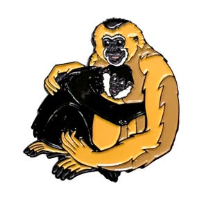 Op de afbeelding: Speld met een moedergibbon die haar baby omhelst. De speld heeft een geel en zwart kleurenschema met een gedetailleerd ontwerp. De moedergibbon heeft een glimlachende uitdrukking en de babygibbon is in haar armen genesteld.