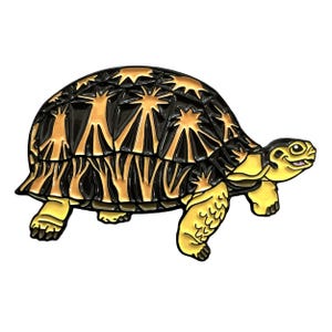 May include: A detailed enamel pin shaped like a tortoise. The tortoise has a black and gold shell with a starburst pattern. The head, legs, and underside are yellow. The tortoise is smiling.