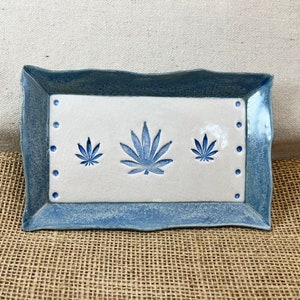 May include: A rectangular ceramic tray with a blue border and a white center. The center features three blue cannabis leaf designs. Small blue dots are placed along the border. The tray has a rustic, handmade appearance.