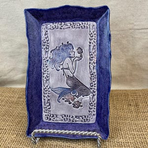 May include: A rectangular ceramic tray with a mermaid design. The tray has a dark blue, textured border and a light gray center. The mermaid has blue hair and a scaled tail. The tray is supported by a silver metal stand.