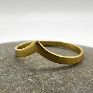 Könnte beinhalten: Ein goldfarbener Ring mit Chevron-Design. Der Ring liegt auf einer grauen Steinoberfläche. Das Band des Rings ist dünn, und die Chevron-Form befindet sich oben.