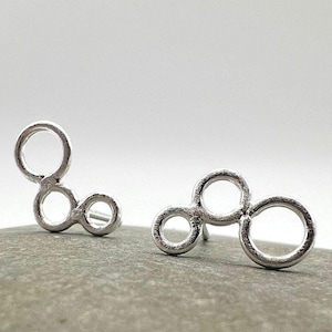 May include: A pair of silver-colored stud earrings. Each earring features a cluster of connected circles, creating a modern, abstract design. The earrings are displayed on a gray stone surface against a white background.