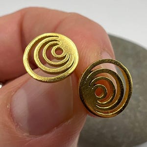 Può includere: Due orecchini a lobo color oro. Ogni orecchino presenta un design circolare con un motivo a spirale. Gli orecchini sono rotondi e hanno una finitura lucida. Sono mostrati su uno sfondo neutro.