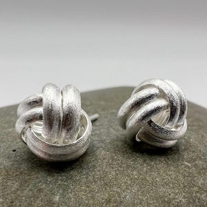 May include: A pair of silver-colored knot stud earrings. The earrings are made of a textured metal and are shaped like a tied knot. The earrings are displayed on a gray stone surface.